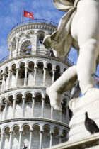 The Campo dei Miracoli or Field of Miracles.The Leaning Tower or Torre Pendente belltower with tourists and a red flag with a white cross the symbol of Pisa under a blue sky and a statue with cherubs...