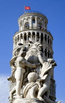 The Campo dei Miracoli or Field of Miracles.The Leaning Tower or Torre Pendente belltower topped with a red flag with a white cross the symbol of Pisa with tourists under a blue sky and a statue with...