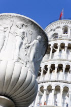 Campo dei Miracoli or Field of Miracles Urn or vase with carving of classical male and female semi naked characters in front of the Leaning Tower belltower or Torre Pendente with tourists walking on a...