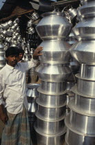 Vendor standing beside stack of vessels at tinware stall in market.Asia Asian Bangladeshi Dacca One individual Solo Lone Solitary Asia Asian Bangladeshi Dacca One individual Solo Lone Solitary