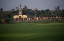 View over rice fields towards crowds gathered around old Hindu temple at sunset. Asia Asian Bangladeshi Farming Agraian Agricultural Growing Husbandry  Land Producing Raising Religion Religious Hindu...