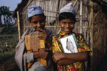 Three-quarter portrait of two  smiling young boys carrying books and writing board standing outside thatched hut.2 Asia Asian Bangladeshi Happy Immature Kids Contented Learning Lessons Teaching Young...