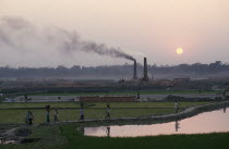 Labourers returning to brick kiln through paddy fields with chimneys releasing trail of thick smoke.air pollution Asia Asian Bangladeshi Ecology Entorno Environmental Environnement Farming Agraian Ag...