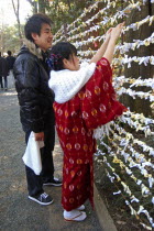Jingumae - at Meijijingu shrine  couple boy and girl early twenty years old  girl in traditional kimono and wrap  boy in down jacket  jeans  sneakers  tie omikuji New years prediction to a fencr.Asia...