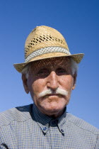 Maditos. Head and shoulders portrait of a Greek old farmer wearing straw hat  looking directly at the lens under strong sunlight and blue sky as background.Face portraitElderlyThird AgeVillage peo...
