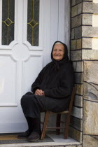 Ano Vrasna. Greek old woman dressed in black and wearing black headscarf is sitting on a chair at the doorstep of her house in front of the houses white door.ElderlyThird AgeVillage peopleTraditio...