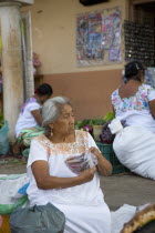 Portrait of an old Mexican saleswoman wearing Yucatan areas traditional white dress with colored flowers while sitting down on the pavement and selling things outside Valladolid citys main market.Foo...
