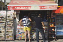 Turkish man and tourist customer at newspaper kiosk also selling bottled water  snacks and cigarettes in afternoon sunshine.Turkish Aegean coastmediterraneanresortformerly HalicarnassusHalikarnas...