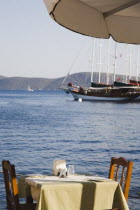 Gulet cruise boat on flat calm water with table  chairs and sun parasol at waterside restaurant in foreground.Turkish Aegean coastmediterraneanresortformerly HalicarnassusHalikarnasregion of Car...