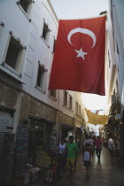 Large Turkish flag hanging between buildings over narrow street in the Old Town bazaar area with crowds passing open shop fronts below.Turkish Aegean coastmediterraneanresortformerly Halicarnassus...