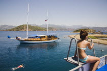 Young Turkish woman in bikini sitting on diving platform on boat off Gemiler or St Nicholas Island with another woman swimming in water below and moored boat beyond.Fethiyeformerly Telmessosformerl...