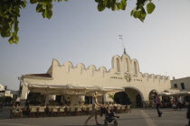 Plateia Eleftherias. Young mother with child in stroller walks through Kos Town Square with view to cafe and market entrance.Greek Islands Resort Summer Clear Blue Sky early season AegeanDestination Destinations Ellada European Greek Southern Europe Bar Bistro Children Immature Kids Mum Restaurant