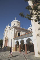 Agia Paraskevi Greek Orthodox Church.  Cream and white facade with colonnaded entrance.  Young woman crossing paved area in foreground.Greek IslandsAegeanMediterraneanKossummerseasonholidayDes...