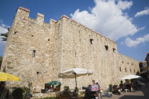 Crenellated exterior walls of stone building with line of outside cafe tables under sun umbrellas in foreground.TurkishAegeancoastresortsummersunshineearly summer seasonMediterraneanholidayD...
