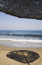 Shadow cast by woven sun umbrella on sandy beach with breaking waves beyond.TurkishAegeancoastresortsummersunshineearly summer seasonMediterraneanholidayDestination Destinations Ellada Europ...