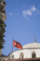 Turkish flag flying in front of white painted mosque with domed roof against blue sky and drifting white cloud.TurkishAegeancoastresortsummersunshineearly summer seasonMediterraneanholidayDe...