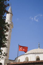 Turkish flag flying in front of white painted mosque with domed roof and minaret against blue sky and drifting white cloud.TurkishAegeancoastresortsummersunshineearly summer seasonMediterranea...