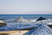 Woven sun umbrellas on empty beach with waves breaking beyond.seasurfTurkishAegeancoastresortSummersunshineearly Summer seasonholidaydestinationdestinations ElladaEuropeanSouthern Europe...
