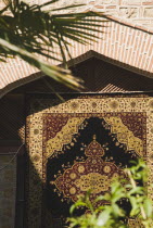 Detail of intricately woven carpet hanging from wall framed by brick portico with palm frond and other leaves in foreground.TurkishAegeancoastresortSummersunshineearly Summer seasonholidaydes...