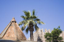 Pointed rooftop and part view of crenellated wall with palm trees against clear  blue sky.TurkishAegeancoastresortSummersunshineearly Summer seasonholidaydestinationdestinations ElladaEurop...