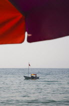 Small boat on water seen from beneath bright red sun umbrella on shore.TurkishAegeancoastresortSummersunshineearly Summer seasonholidaydestinationdestinations ElladaEuropeanSouthern Europe...