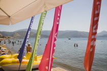 Marmaris beach with water sports equipment including line of yellow pedelos for hire and brightly coloured banners advertising services.  Tourists in calm sea and sunbathing in background with distant...