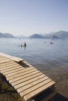 Wooden jetty for local ferry boat in foreground with people swimming and standing in flat calm water with distant coastline beyond.Turkish RivieraresortholidayDestination Destinations Ellada Europ...