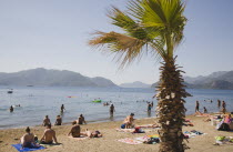 Marmaris on the Turkish Riviera in early Summer season.   Turkish holidaymakers sunbathing on town public beach in afternoon sun with palm tree in foreground and view towards coastline beyond.coastc...