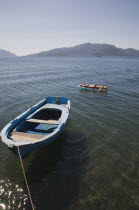 Tanned woman sunbathing on floating lilo on flat  calm sea with blue painted  local fishing boat moored in the foreground in early Summer season.coastcoastalresortsunbedsuntansunshinestillholi...