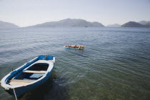 Tanned woman sunbathing on floating lilo on flat  calm sea with blue painted  local fishing boat moored in the foreground in early Summer season.coastcoastalresortsunbedsuntansunshinestillholi...