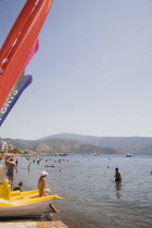 Marmaris beach on the Turkish Riviera.  Young female tourist in bikini and baseball cap at waters edge sitting on yellow pedalo.  Water sports equipment for hire with brightly coloured flags advertisi...