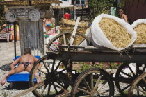 European male tourist sunbathing on lounger in hotel resort garden with display of traditional Turkish farm equipment  cart and straw and ancient Greek column used for sun shade support.coastcoastal...