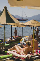 Female tourists sunbathing on hire sunbeds with parasols on Marmaris beach with Gulet on calm sea beyond in late afternoon sun in early Summer season.Turkish Rivieracoastcoastalresortsunshinesun...