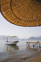 Local water taxi moored at wooden jetty from shade of part seen straw parasol in early Summer sun with calm  flat water.Turkish Rivieracoastcoastalresortsunshinesun umbrellaholidaydestination...