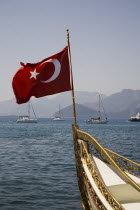 Turkish flag flying from prow of decorated water taxi with Gulet and yachts in the background on calm  flat sea.formerly Greek Physkos in CariaTurkish RivieracoastcoastalOttomanresortholidaySu...