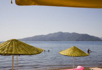 Parasols and sunbeds for hire on beach with views over bay on clear bright early Summer day.  Man emerging from calm clear water with heads of other swimmers seen behind.formerly Greek Physkos in Car...