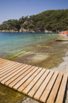 Olu Deniz.  Slatted wooden jetty for pleasure boats on beach in foreground with view towards tree covered coast behind and people swimming in aquamarine water.Turkish rivieraAegeancoastcoastalOlu...