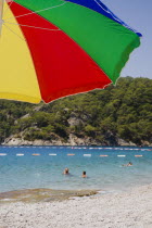 Olu Deniz.  Tourists swim at entrance to lagoon and beach rated in top five worldwide.  Part seen brightly coloured striped parasol in foreground.Turkish RivieraAegeancoastcoastalOludenizseashorecalmclearshallowazurebrightsunSummerresortholidayholidaymakersswim5 Colored Destination Destinations European Middle East Salt Water Waves Sand Sandy Beaches Tourism Seaside Shore Tourist Tourists Vacation Sunbather Travel Turkiye Western Asia