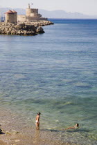 Rhodes Town.  View from St Pauls Gate entrance to Old Town across clear turquoise sea towards stone windmills and fortified tower with tourists bathing in shallow water in foreground.Aegeancoastcoa...