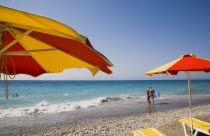 Red and yellow striped parasols and yellow and white sun loungers on Ixia beach resort looking north towards Turkish coast.  Tourists swimming and standing at edge of clear  aquamarine sea and breakin...