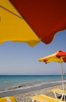 Red and yellow striped parasols and yellow and white sun loungers on Ixia beach resort looking north towards Turkish coast.  Tourists swimming in clear  aquamarine sea with breaking surf.Aegeancoast...