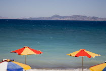 Orange  yellow and blue striped tops of sun umbrellas against expanse of aquamarine sea and view to distant coastline. GreekAegeanCoast - CoastalRodiPackage holidaydestination resortsun sunshin...