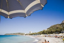 Ixia beach.  View east from beneath partly seen sun umbrella towards Rhodes town with tourists on line of hire sunbeds along Ixia bay on cloudless Summer morning.AegeanRodiseacoast coastalpackage...