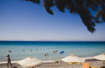 Ixia beach.  Line of sun umbrellas and sunbeds with tourists swimming in clear shallow aquamarine water.  Overhanging palm fronds in foreground. AegeanRodiseacoast coastalpackage holiday resorts...