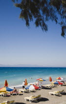 Ixia beach.  Line of brightly coloured  striped sun umbrellas and sunbeds with tourists sunbathing and swimming in clear shallow aquamarine water. AegeanRodiseacoast coastalpackage holiday resort...