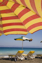 Ixia beach resort popular for water sports with red and yellow striped sun umbrella in foreground against blue  cloudless sky.  Sun loungers and parasol overlooking sea beyond.AegeanRodiseacoast c...