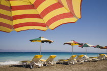 Ixia beach resort popular for water sports with part seen red and yellow striped sun umbrella in foreground against blue  cloudless sky.  Line of sun loungers and parasols overlooking sea beyond.Aege...