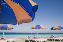 Ixia beach resort popular for water sports with line of blue and orange striped sun umbrellas and sun loungers in foreground against blue  cloudless sky and sea view with distant windsurfer behind.Ae...