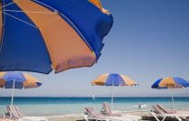 Ixia beach resort popular for water sports with line of blue and orange striped sun umbrellas and sun loungers in foreground against blue  cloudless sky and sea view with distant windsurfer behind.Ae...