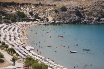 Lindos.  Shallow bay with tourists swimming and small  moored boats in high summer season.  Lines of sun umbrellas and loungers following curve of beach.AegeanseaRodicoast coastalswimbathingpac...